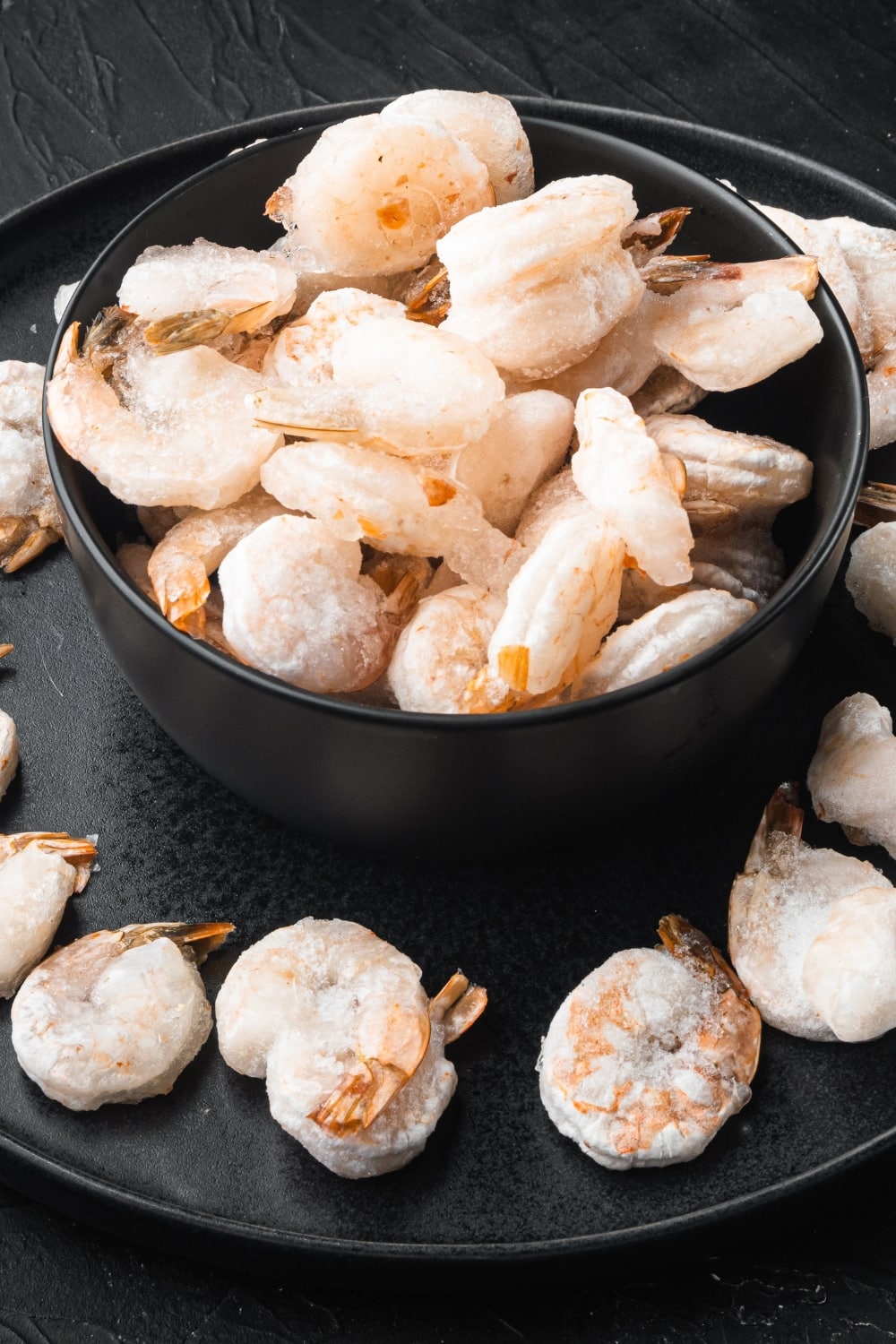 How To Thaw Frozen Shrimp Properly featuring Frozen Shrimp in a Black Bowl on a Black Plate with More Shrimp Around the Bowl