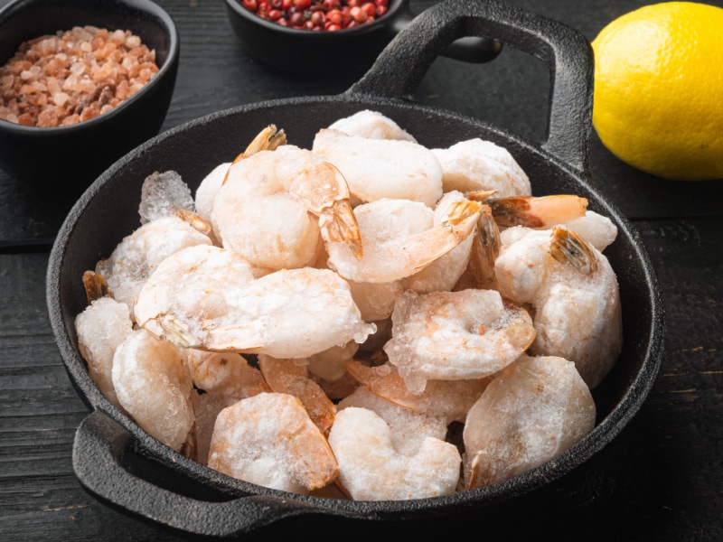 Frozen Shrimp in Cast Iron Pan On Black Wooden Table with Lemon and Seasonings in Background